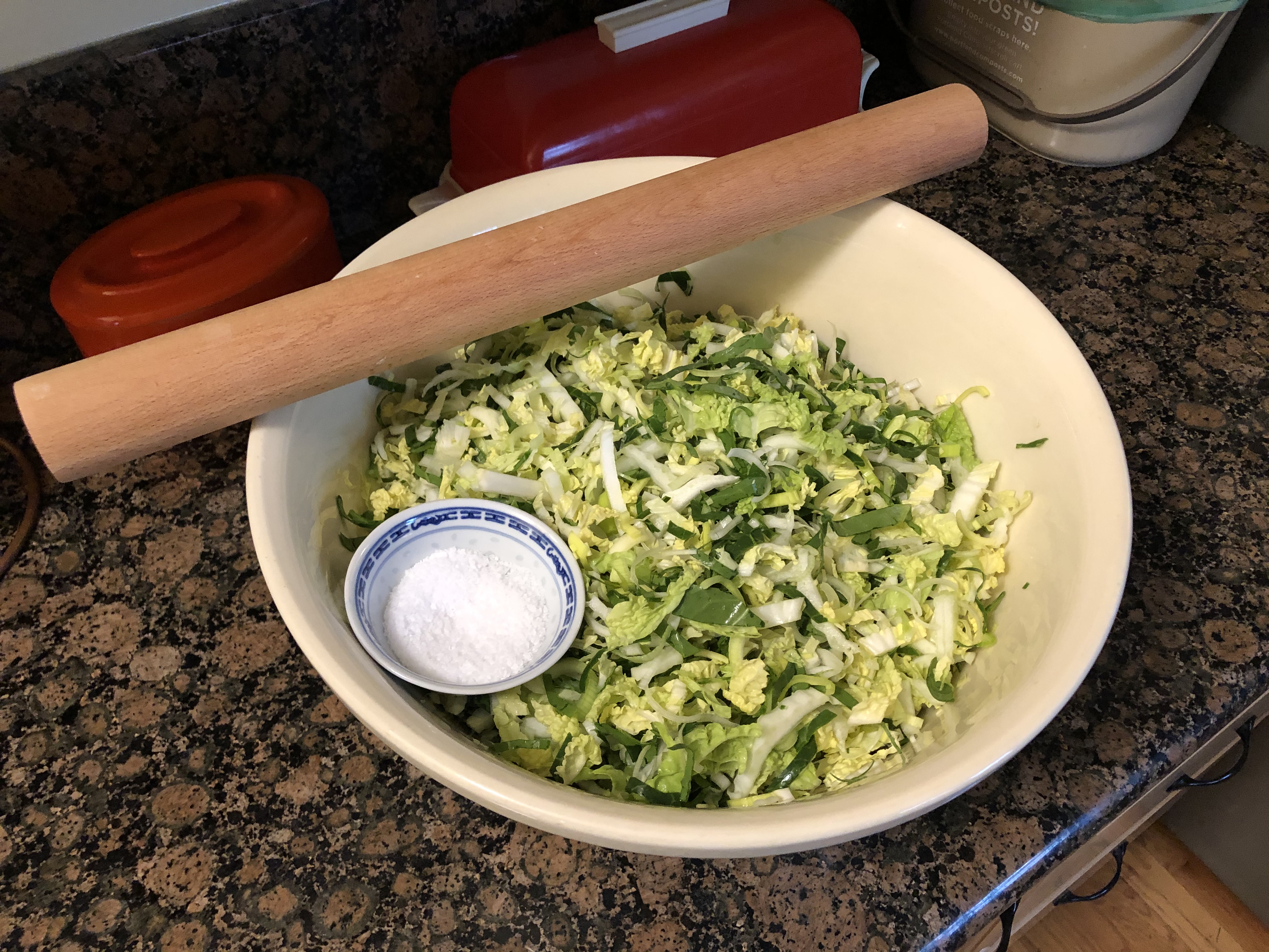 Very large pottery bowl filled with shredded vegetables. Across the top of bowl is a wooden rolling pin for thumping the veg. A small bowl of kosher salt sits to one side.